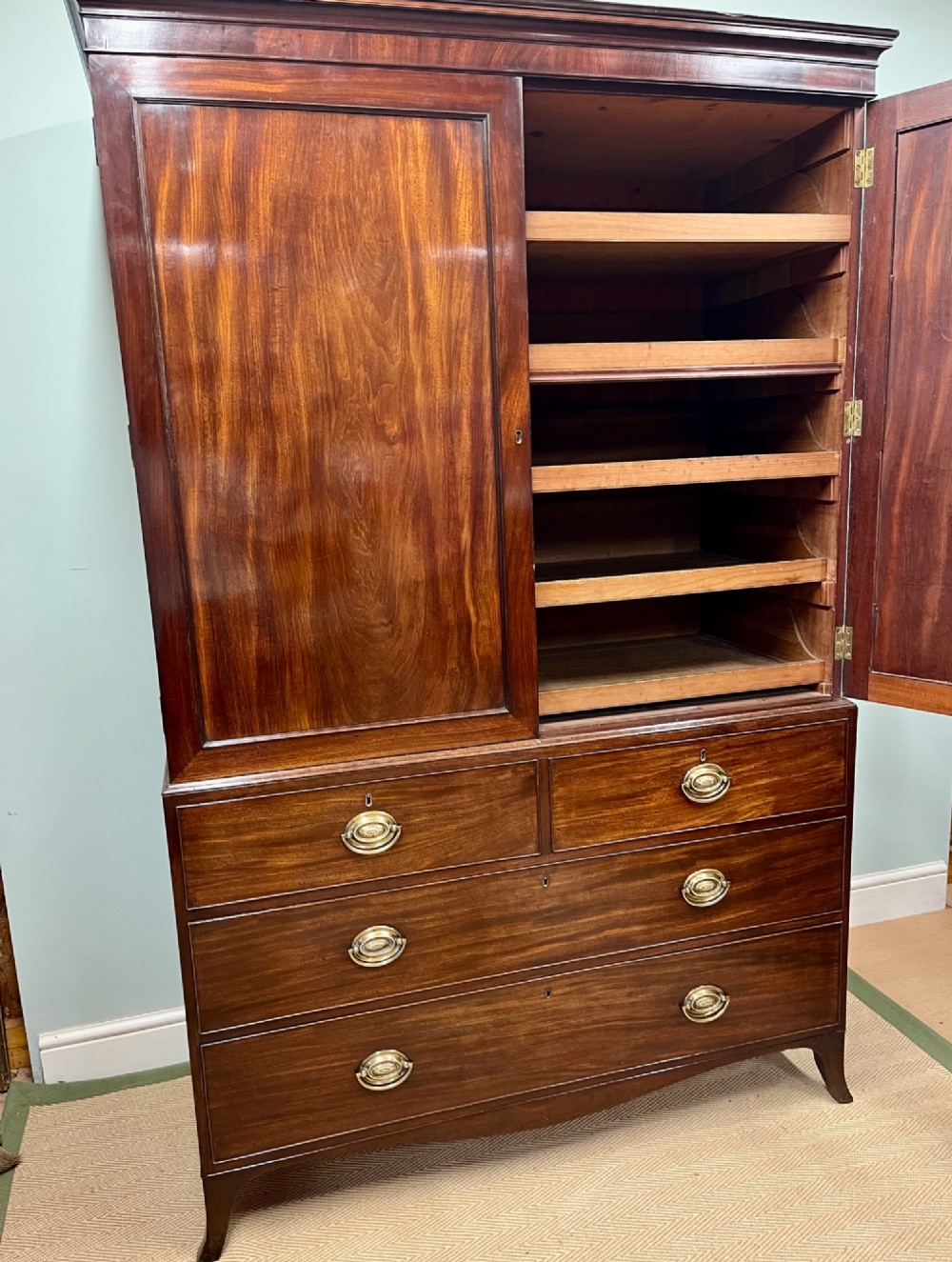 A Regency mahogany linen press featuring an upper two-door cupboard with flame veneer panels above a base of two short and two long drawers with brass handles. View IV