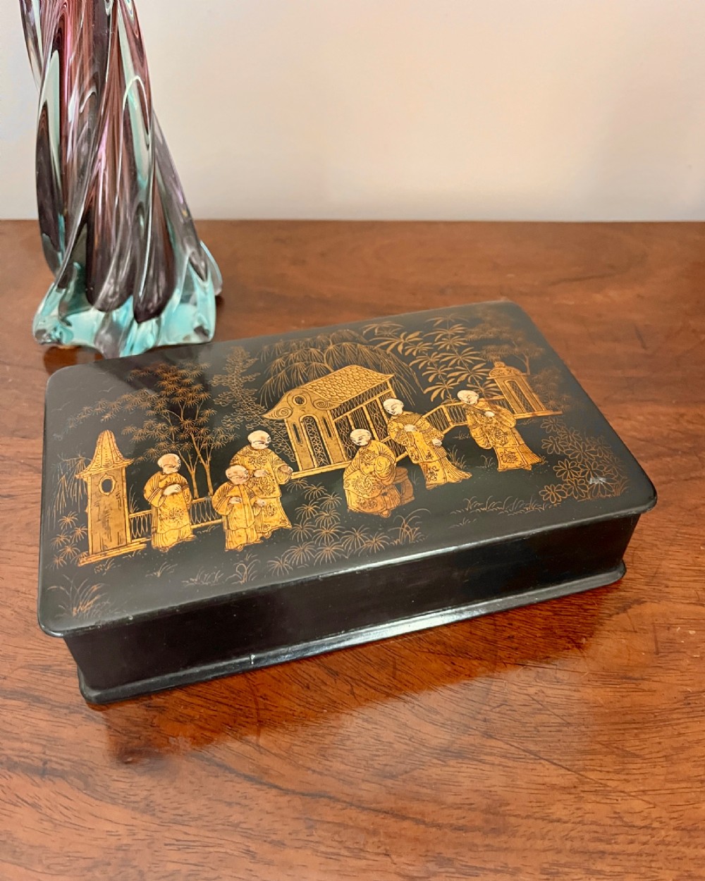 A rectangular black lacquered box decorated with detailed gold Chinoiserie scenes depicting figures, foliage, and pavilions. View V