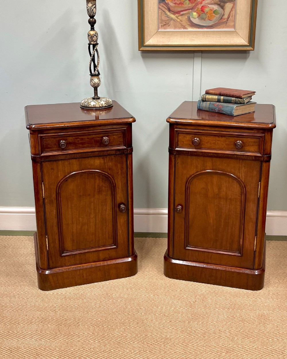 A matching pair of 19th-century polished mahogany bedside cupboards featuring top drawers, arched panel doors, and plinth bases.