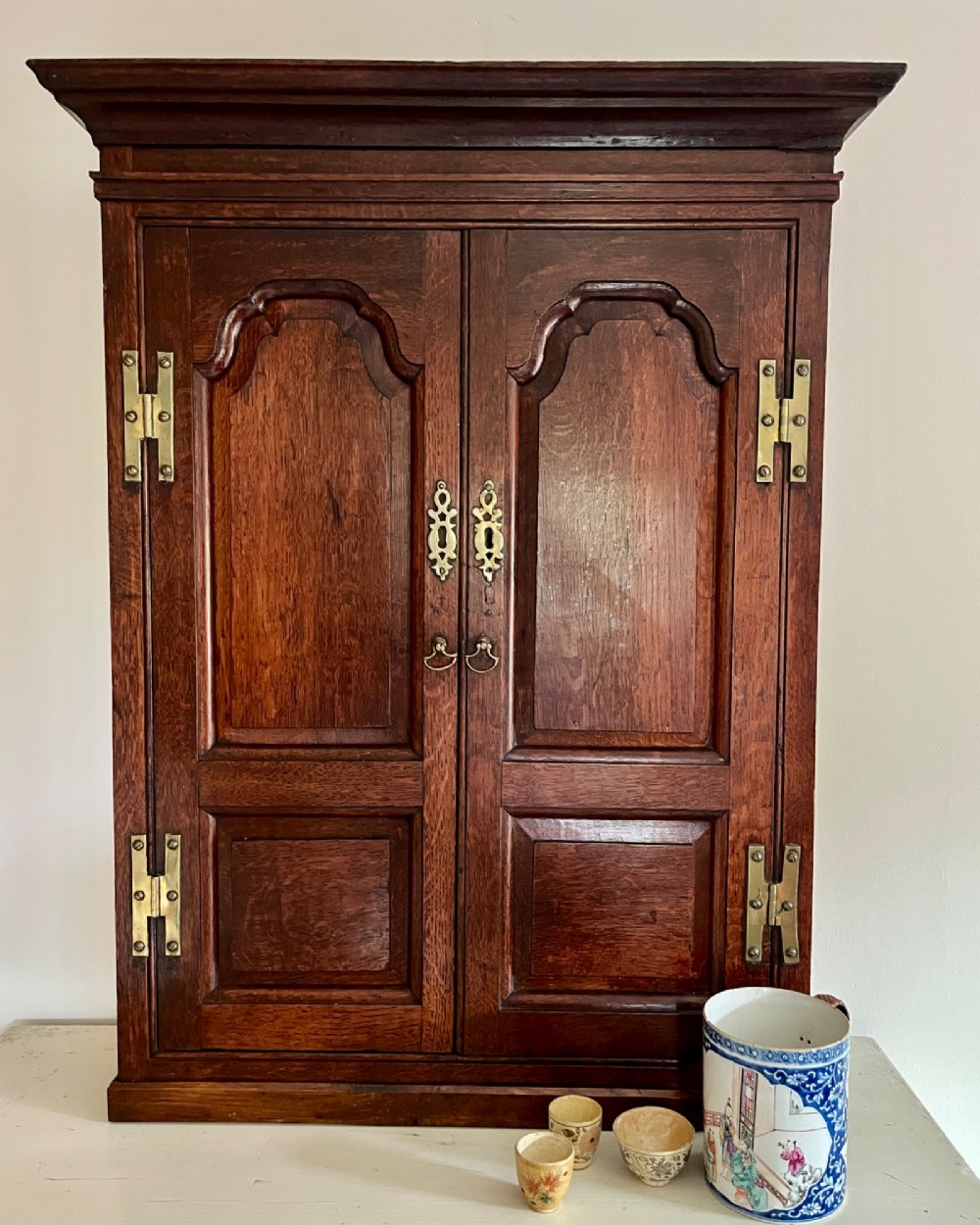 An eighteenth-century dark oak wall cupboard featuring twin arched fielded panel doors and prominent brass H-hinges.