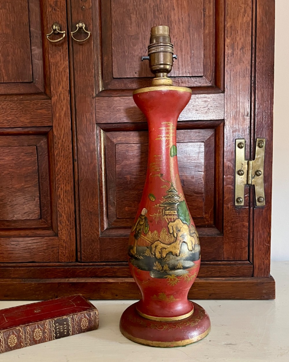 A red lacquered table lamp featuring hand-painted gilt Chinoiserie figures and pagodas on a circular base with a brass bulb socket.