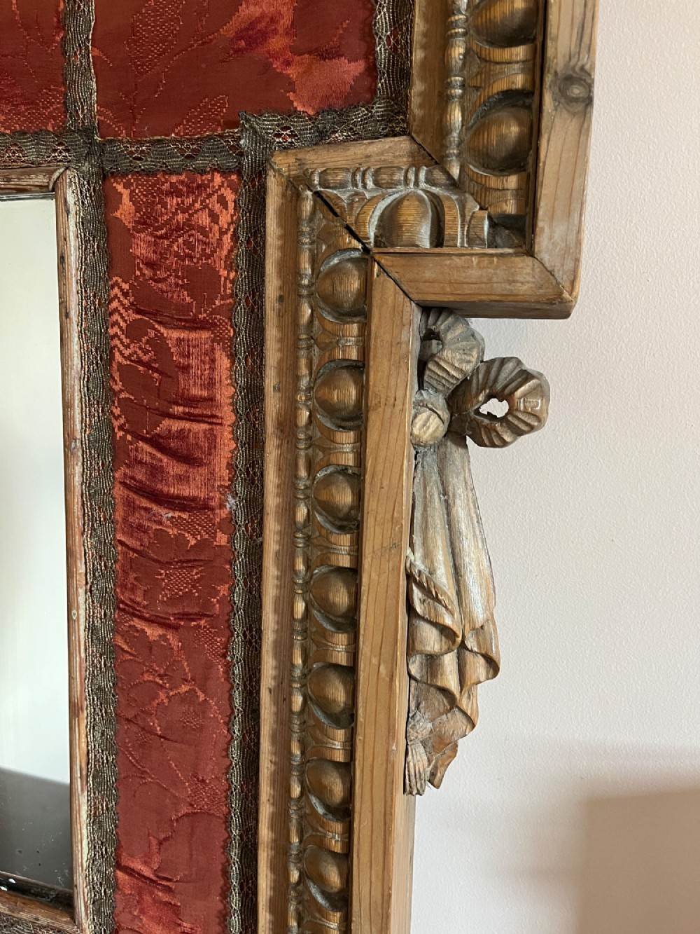 An 18th-century carved pine wall mirror featuring egg-and-dart outer borders, side ear brackets, and a red damask fabric panel surrounding the central rectangular glass plate. View VI