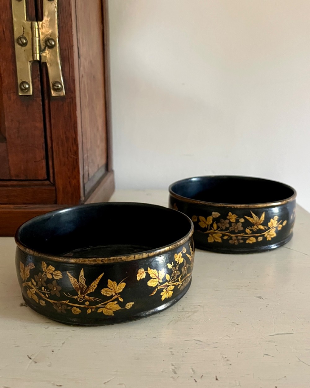 A pair of circular black lacquered papier-mache wine coasters decorated with hand-painted gold botanical and dragonfly motifs.