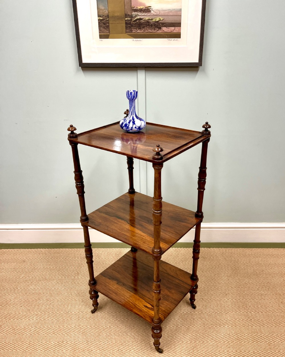 A 19th-century rosewood three-tier etagere featuring turned spindle corner posts, top finials, and brass castors on the feet.