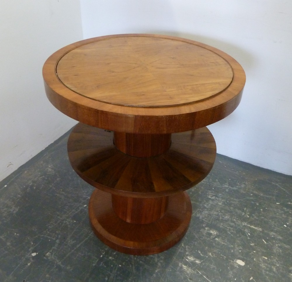 A circular Art Deco walnut metamorphic card table featuring a quarter-veneered top, central cylindrical column, and tiered circular shelf.