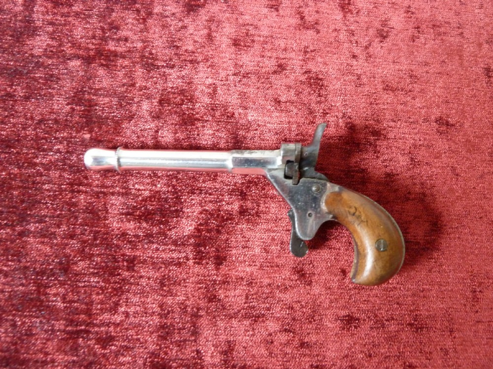 A small 19th-century postmans dog scarer pistol featuring a turned metal barrel, exposed hammer mechanism, and contoured wooden handle. View II