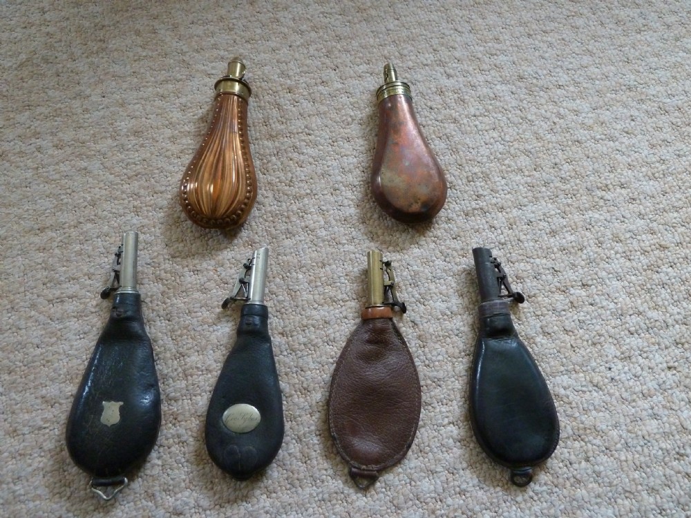 A collection of six 19th-century powder and shot flasks made from embossed copper, brass, and dark brown leather.