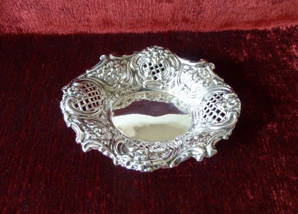 An oval Edwardian sterling silver pin dish featuring pierced lattice panels, repousse floral scroll borders, and a polished centre. View II