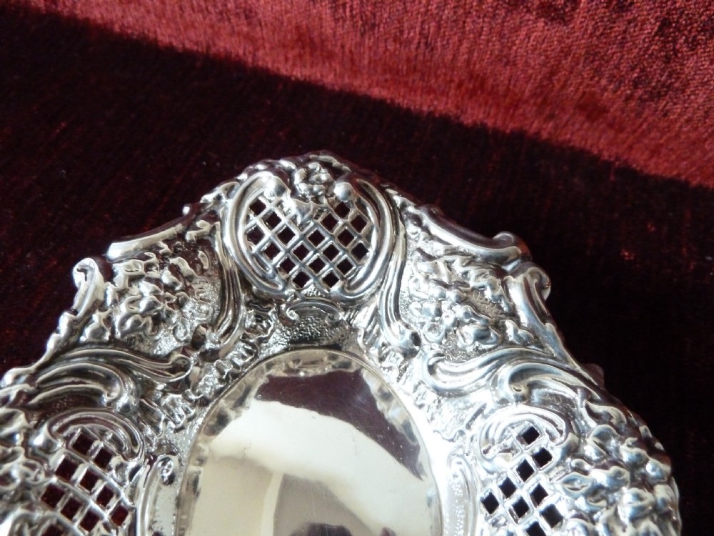 An oval Edwardian sterling silver pin dish featuring pierced lattice panels, repousse floral scroll borders, and a polished centre. View IV