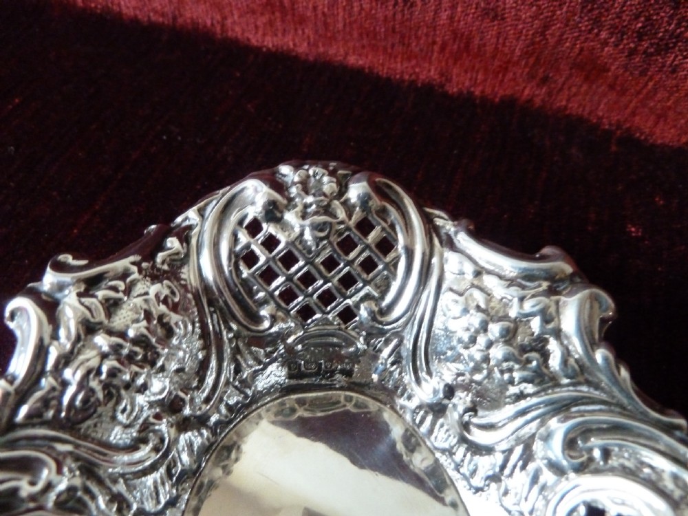 An oval Edwardian sterling silver pin dish featuring pierced lattice panels, repousse floral scroll borders, and a polished centre. View V