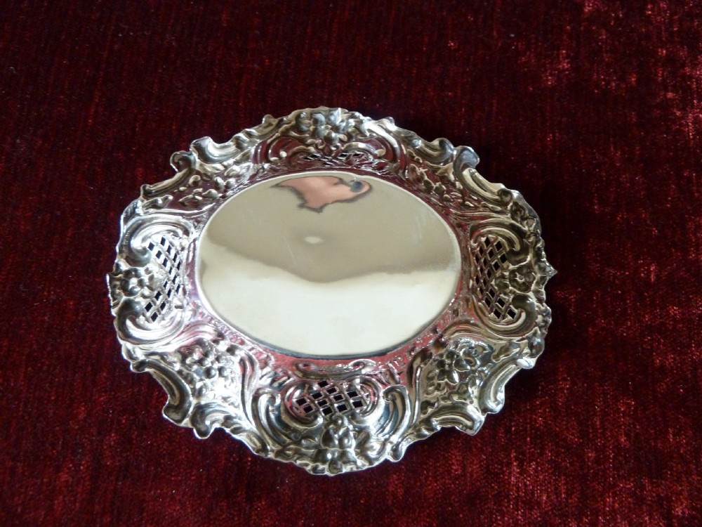 An oval Edwardian sterling silver pin dish featuring pierced lattice panels, repousse floral scroll borders, and a polished centre. View VI