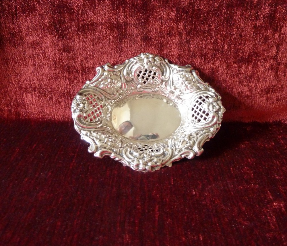 An oval Edwardian sterling silver pin dish featuring pierced lattice panels, repousse floral scroll borders, and a polished centre. View VII