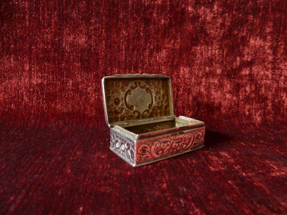 An open rectangular Victorian sterling silver snuff box displaying embossed scrollwork decoration and a gilded interior.