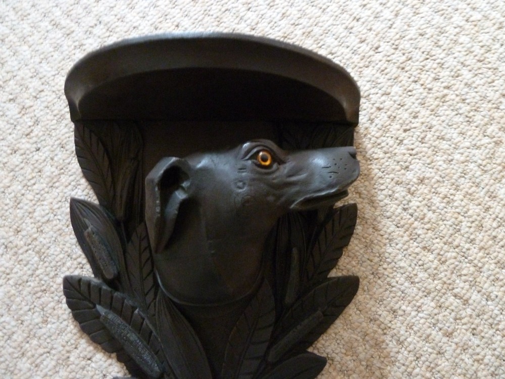 A dark brown hand-carved wooden wall bracket featuring a dog head surrounded by carved leaves below a small rounded shelf. View X