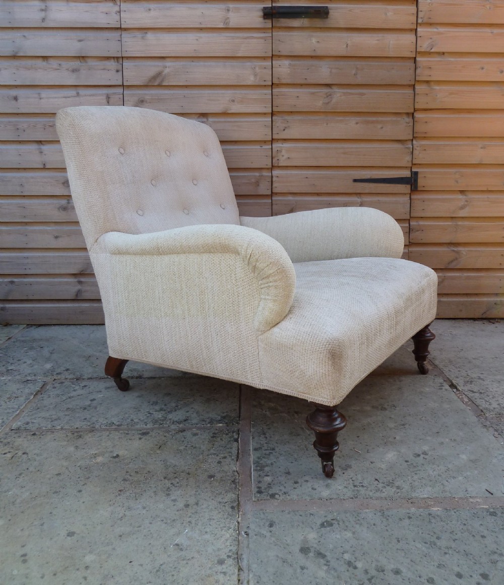 A low-slung, cream fabric upholstered armchair with a buttoned backrest, padded scroll arms, and dark turned legs on castors.
