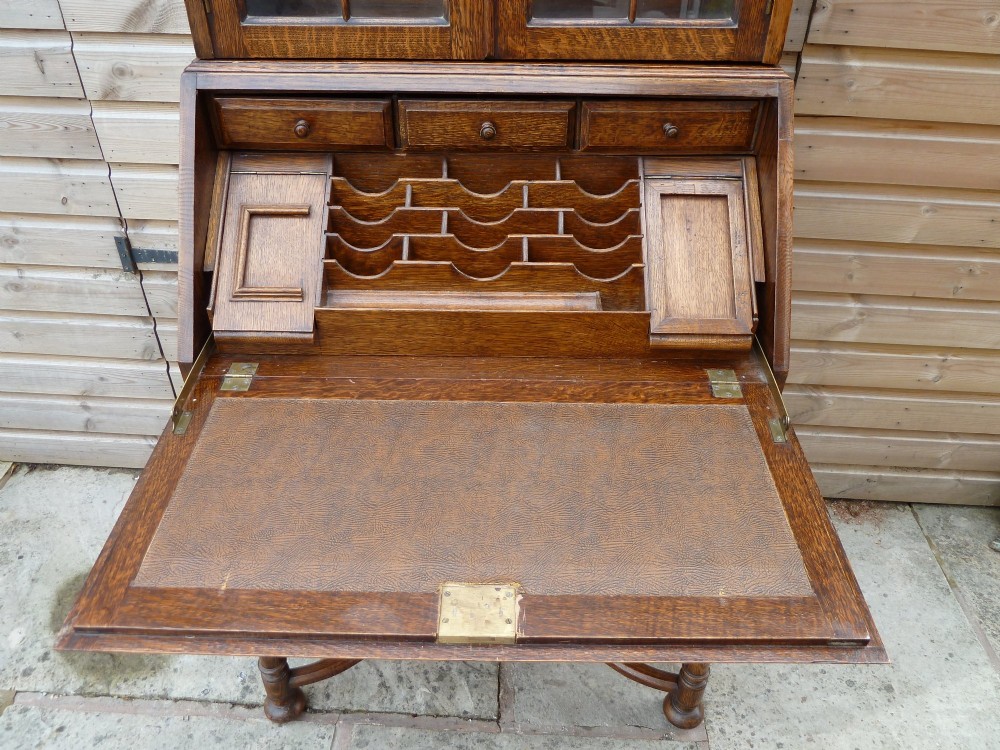 An antique oak bureau bookcase featuring an arched glazed upper cabinet, carved slant-front desk flap, drawers, and turned legs with an X-stretcher. View V