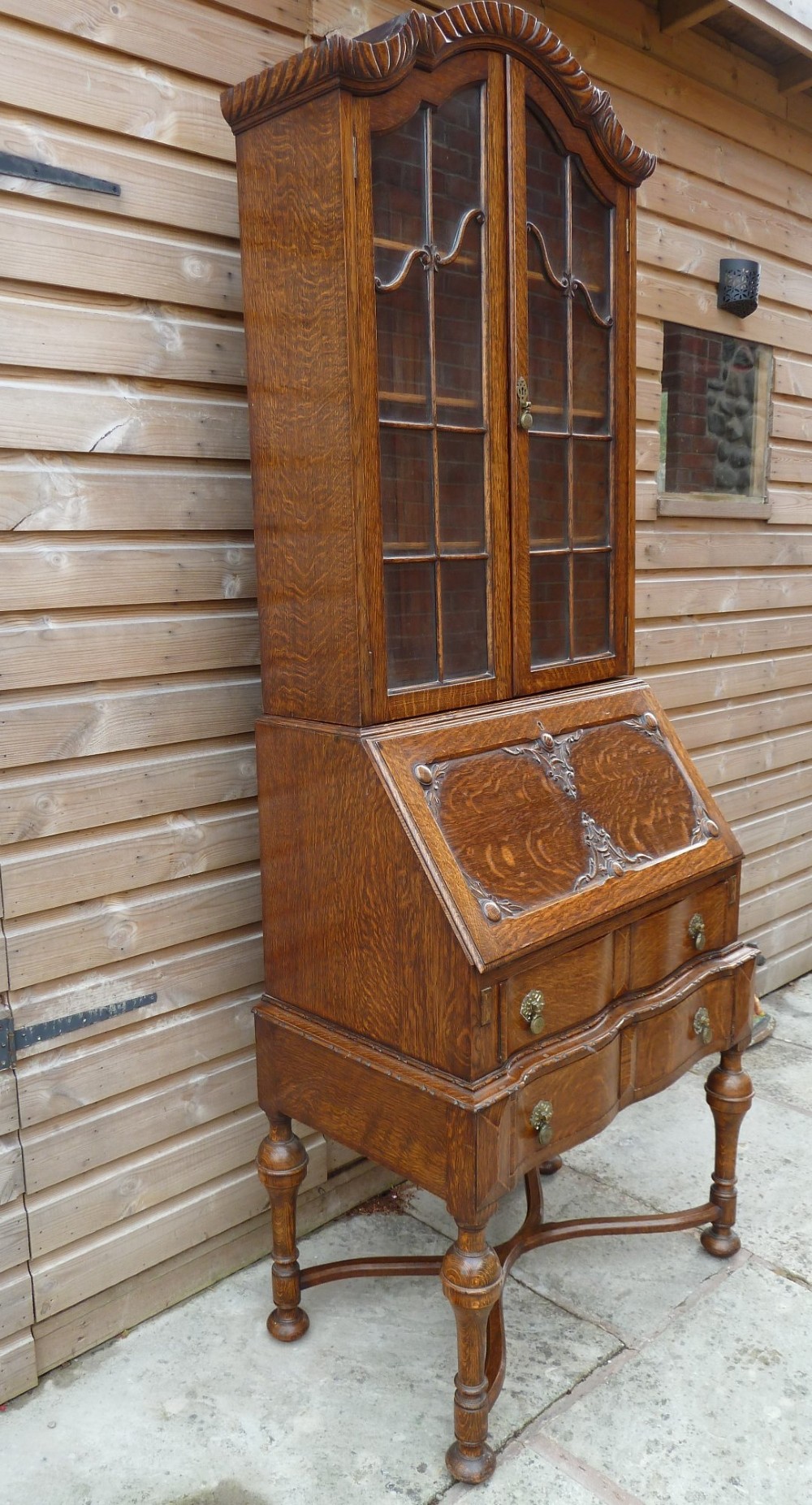 An antique oak bureau bookcase featuring an arched glazed upper cabinet, carved slant-front desk flap, drawers, and turned legs with an X-stretcher. View VI
