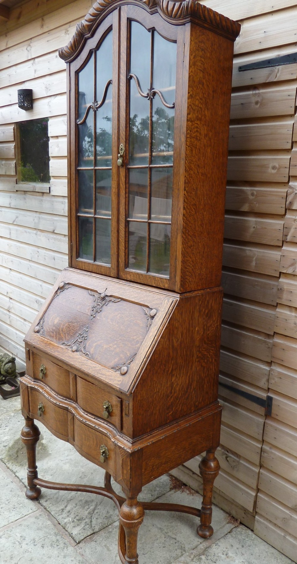 An antique oak bureau bookcase featuring an arched glazed upper cabinet, carved slant-front desk flap, drawers, and turned legs with an X-stretcher. View VII