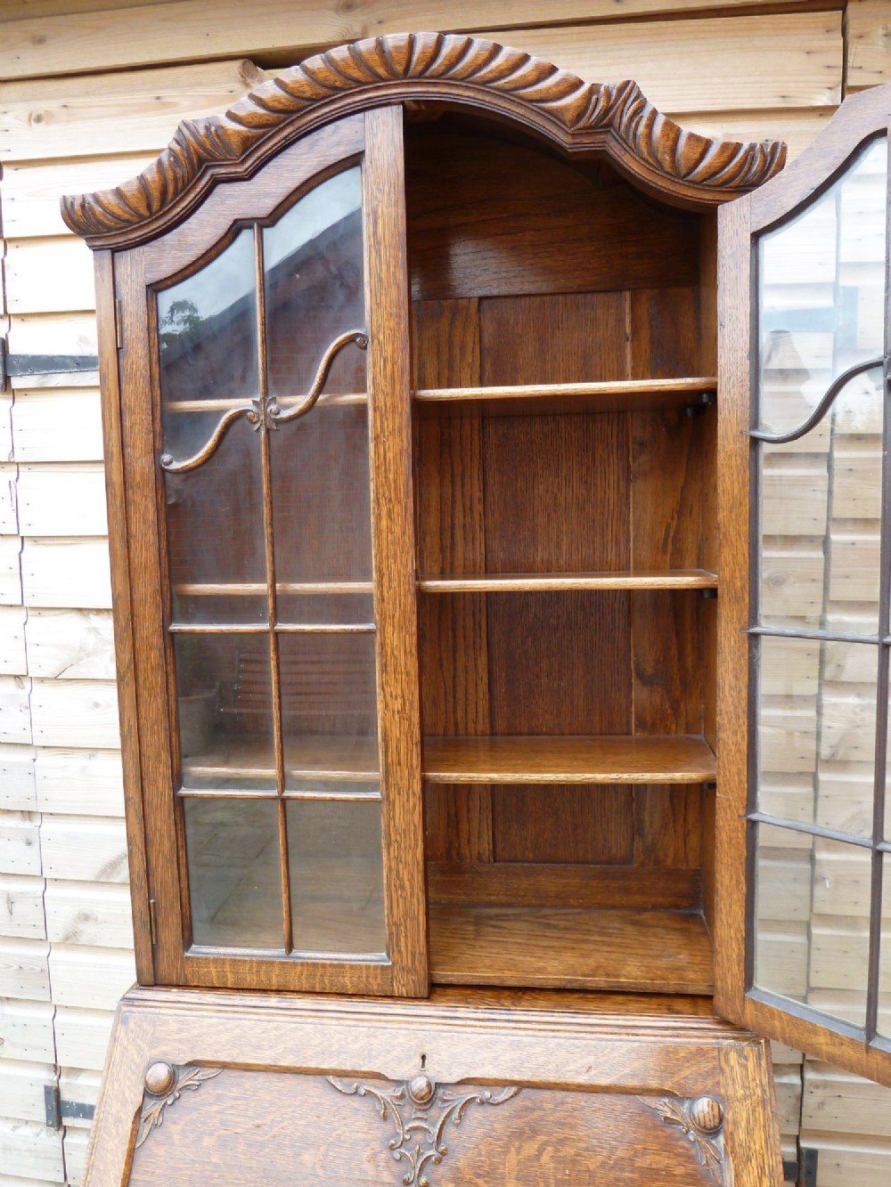 An antique oak bureau bookcase featuring an arched glazed upper cabinet, carved slant-front desk flap, drawers, and turned legs with an X-stretcher. View IX