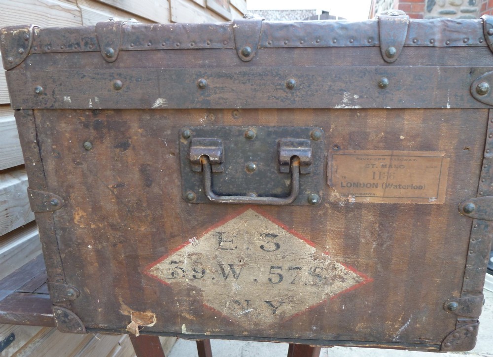 An open 19th-century flat-top steamer trunk featuring a brown striped canvas exterior, brass hardware, wood slatted bindings, and a cream quilted fabric interior lid lining. View V