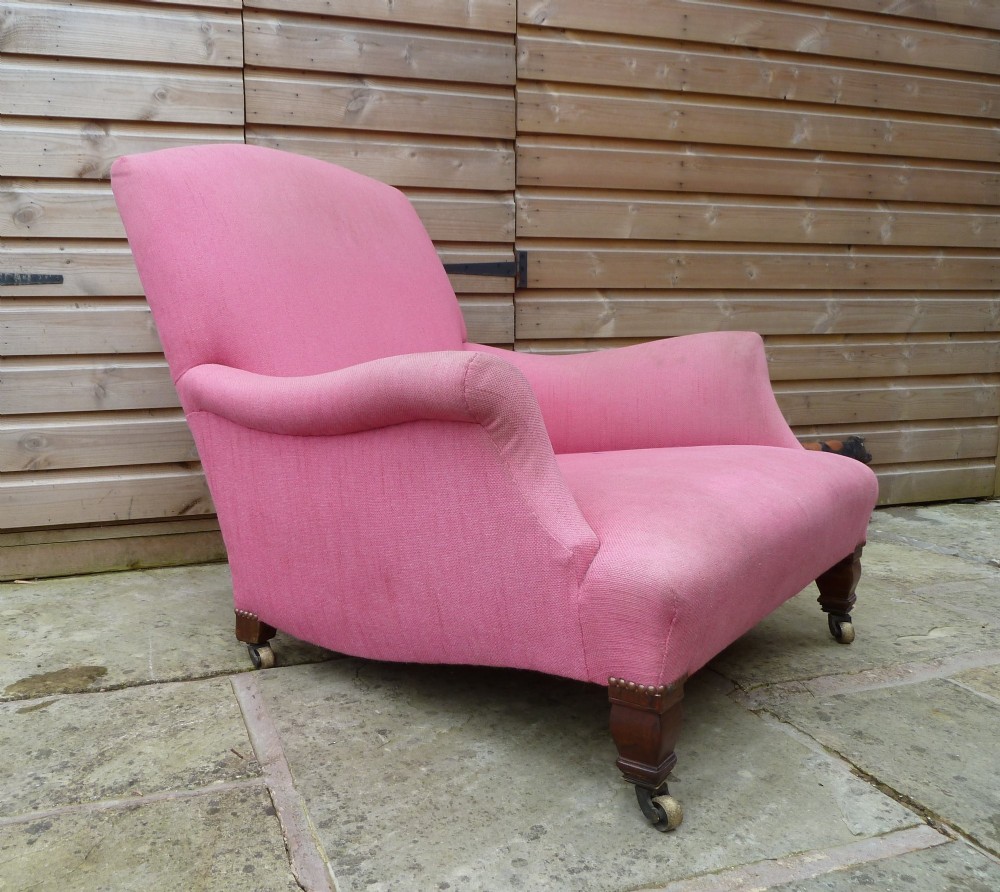 A deep-seated Howard style armchair upholstered in pink fabric, featuring low scrolled arms, turned mahogany front legs, and brass castors.