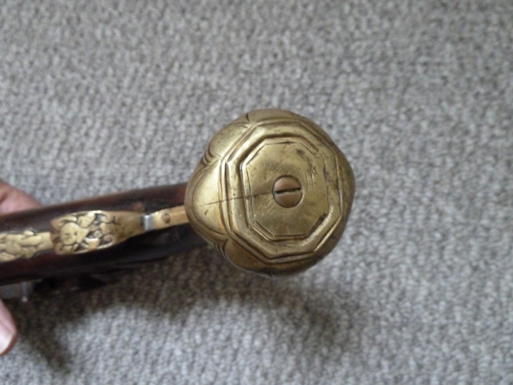 An 18th-century flintlock pistol featuring a carved dark walnut stock, engraved brass butt cap, brass ramrod pipes, and an iron lock mechanism. View IX
