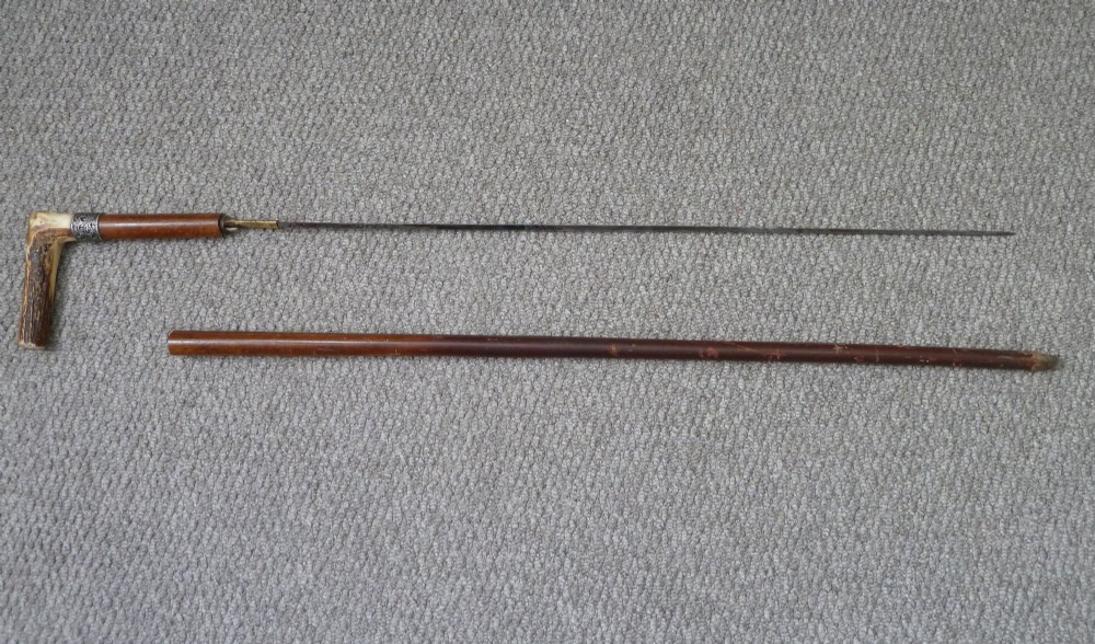 A Victorian sword stick displayed unsheathed in two parts, showcasing a stag horn handle, decorative metal ferrule, dark wooden scabbard, and a narrow steel blade.