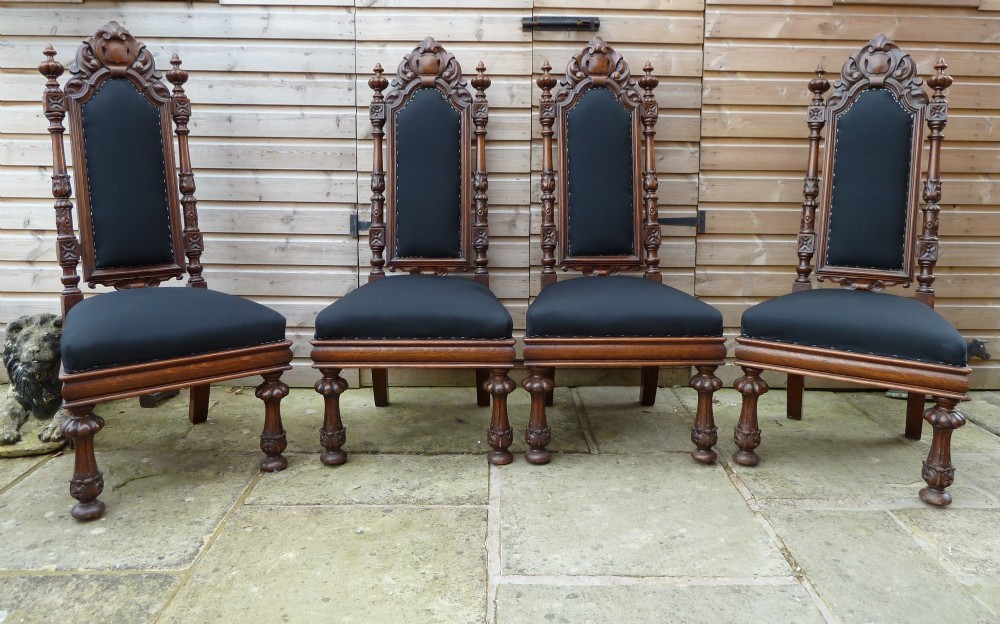 A set of four Victorian Gothic style carved oak high-back chairs featuring black upholstered seats and backrests with ornate cresting and turned legs.