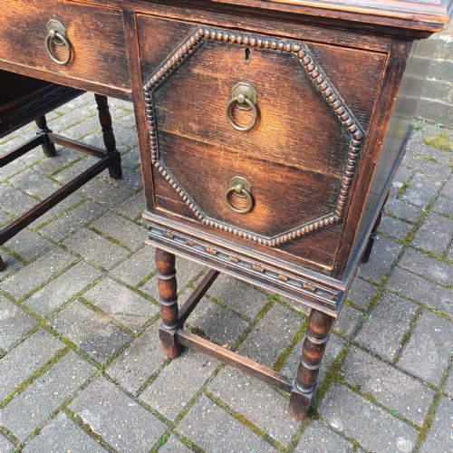 fig 6. A vintage dark oak writing desk with five drawers, geometric bead moulding, turned legs, brass handles, and a black leather inset top.