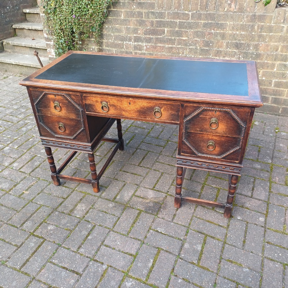 A vintage dark oak writing desk with five drawers, geometric bead moulding, turned legs, brass handles, and a black leather inset top.