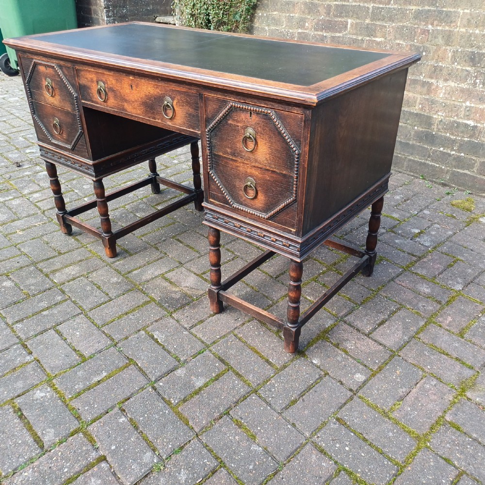 A vintage dark oak writing desk with five drawers, geometric bead moulding, turned legs, brass handles, and a black leather inset top. View VI