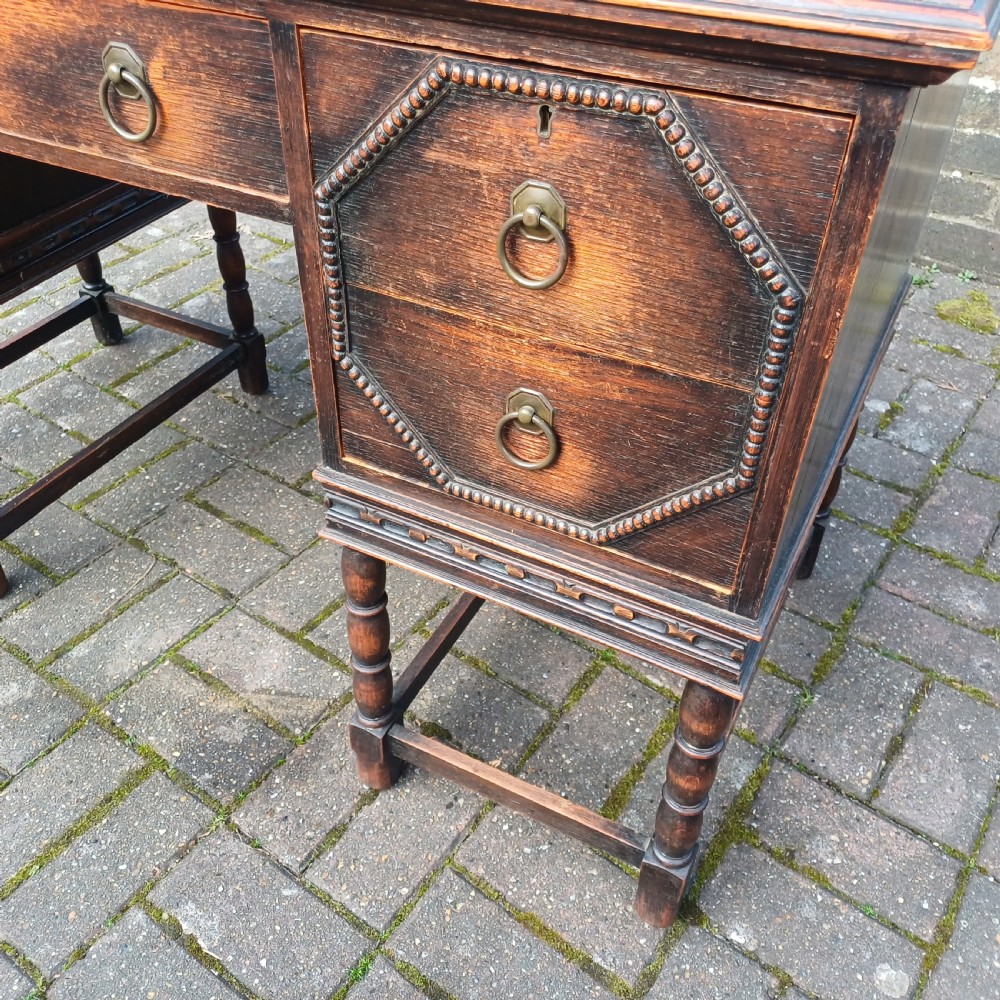 A vintage dark oak writing desk with five drawers, geometric bead moulding, turned legs, brass handles, and a black leather inset top. View VII