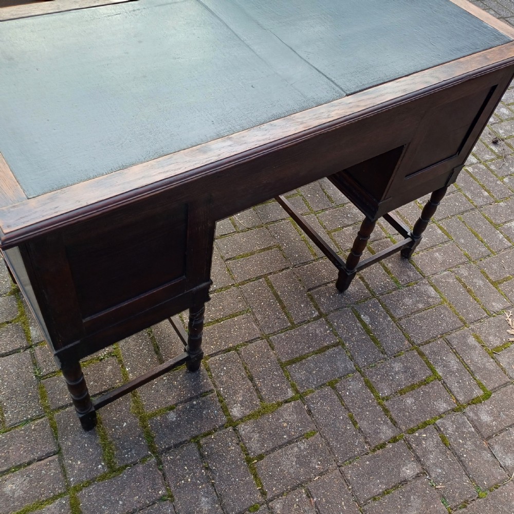 A vintage dark oak writing desk with five drawers, geometric bead moulding, turned legs, brass handles, and a black leather inset top. View X