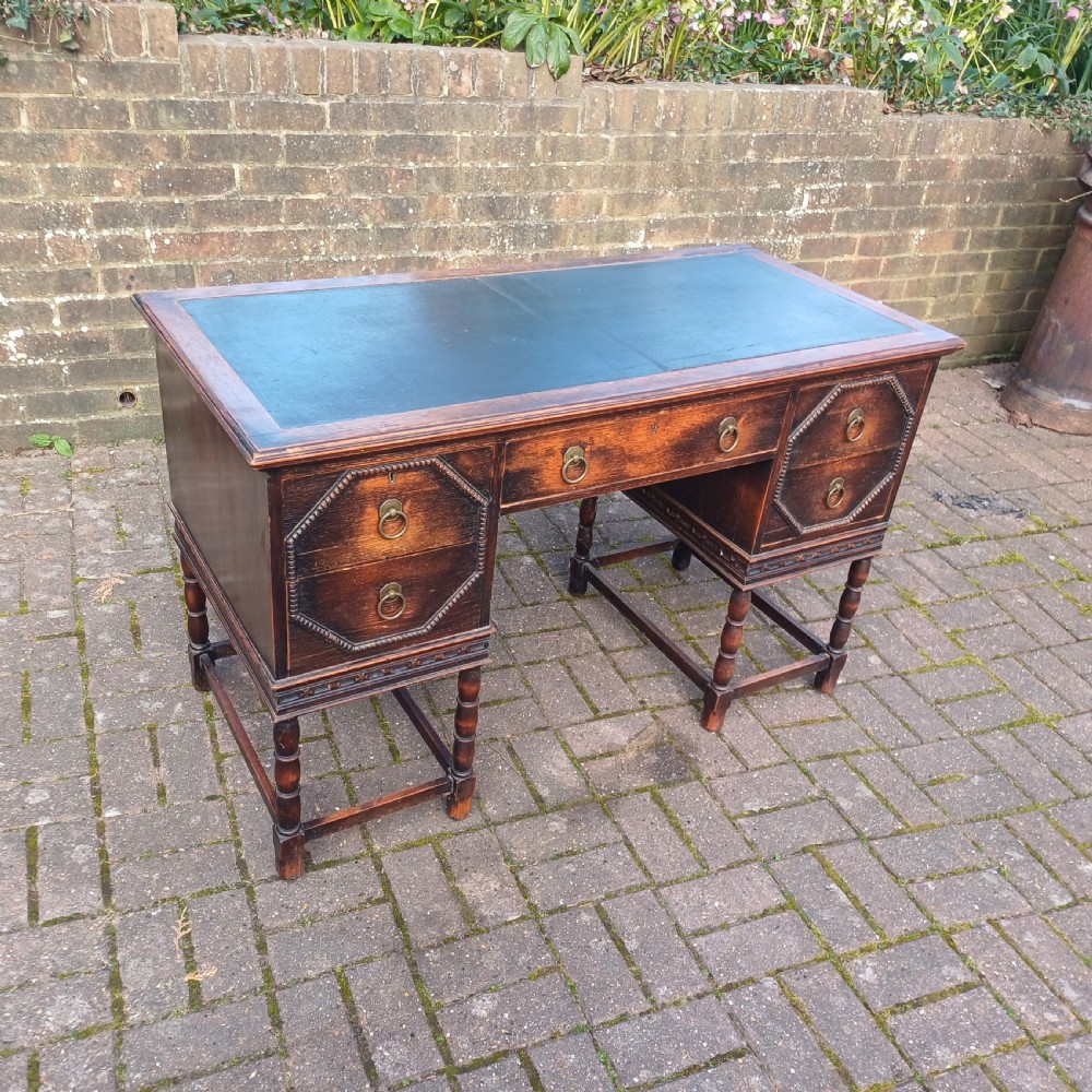 A vintage dark oak writing desk with five drawers, geometric bead moulding, turned legs, brass handles, and a black leather inset top. View XI