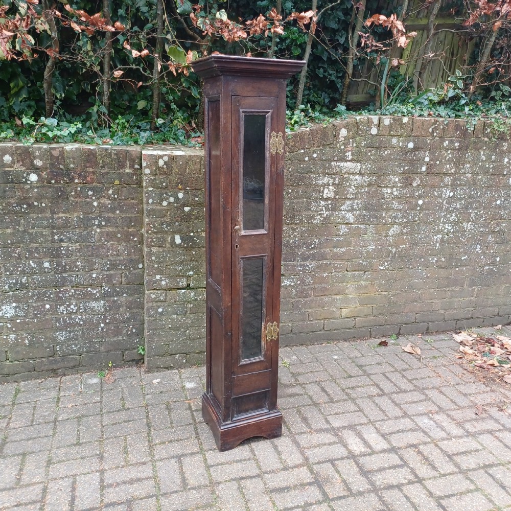 A tall, narrow Victorian dark oak gun cupboard featuring two glass panels set into a single front door with decorative brass hinges. View II