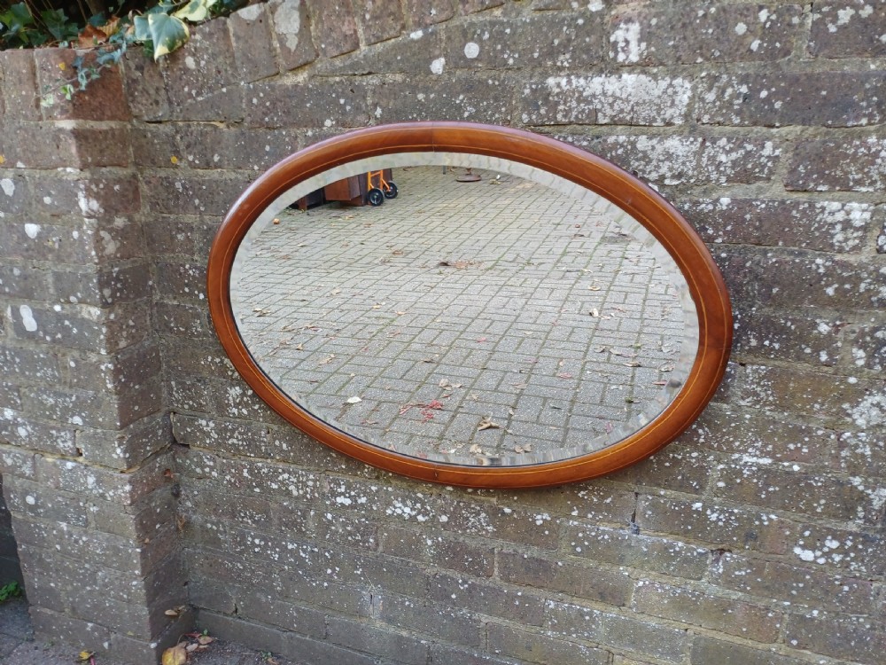 An oval mahogany framed wall mirror featuring a bevelled glass edge and subtle fine string inlay detail around the perimeter.