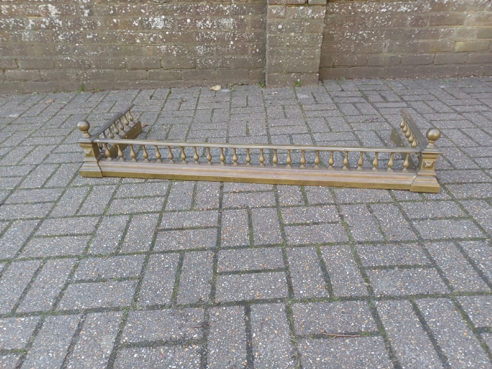 An antique brass fireplace fender featuring a turned spindle balustrade, spherical corner finials, and a stepped plinth base.