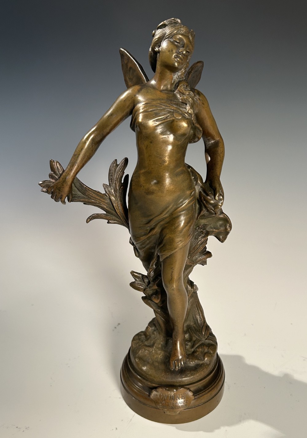 A patinated bronze sculpture of a winged female figure standing on a circular moulded base with an inscribed title plaque.