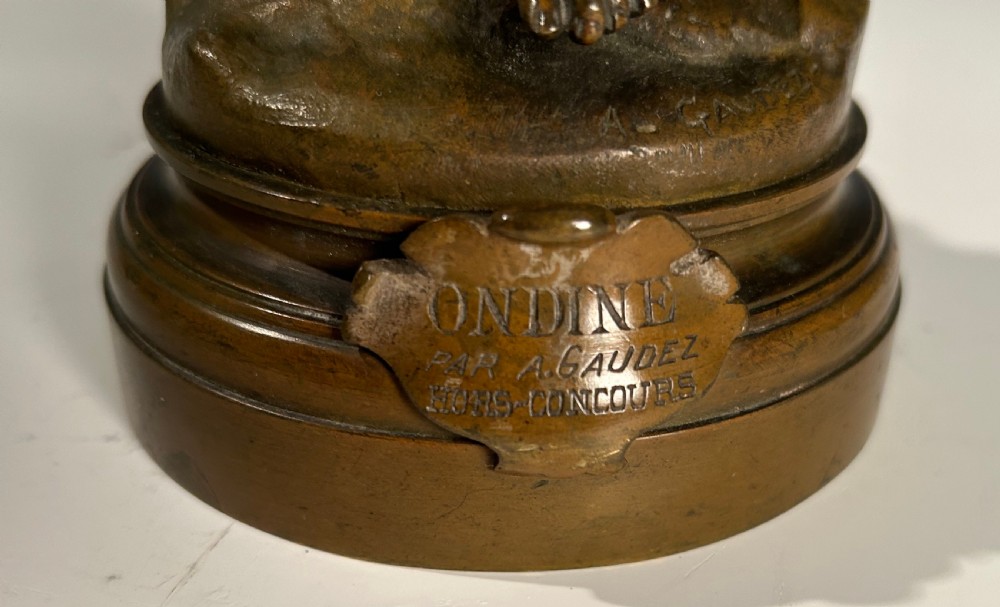 A patinated bronze sculpture of a winged female figure standing on a circular moulded base with an inscribed title plaque. View II