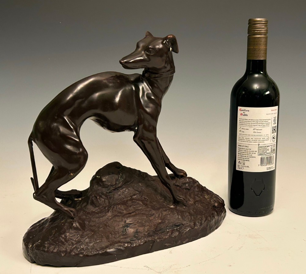 A dark brown patinated bronze sculpture depicting an alert standing greyhound on a textured naturalistic base. View III