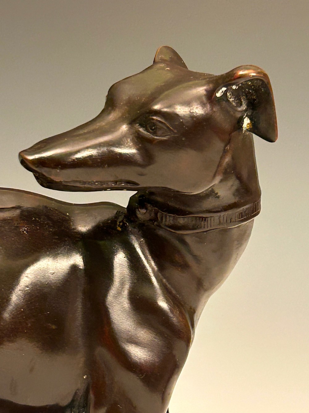 A dark brown patinated bronze sculpture depicting an alert standing greyhound on a textured naturalistic base. View V