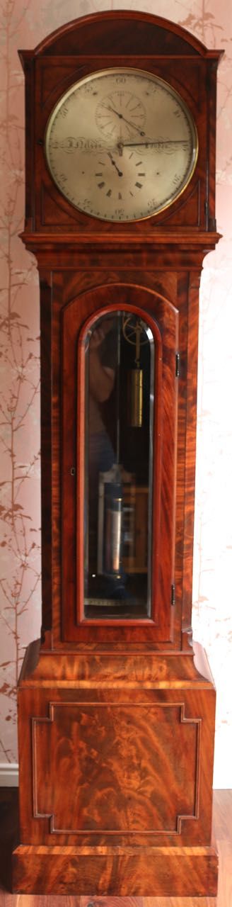 fig 1. A Regency flame mahogany astronomical regulator longcase clock featuring an arched hood, silvered dial with three sub-dials, glass trunk door, and plinth base.