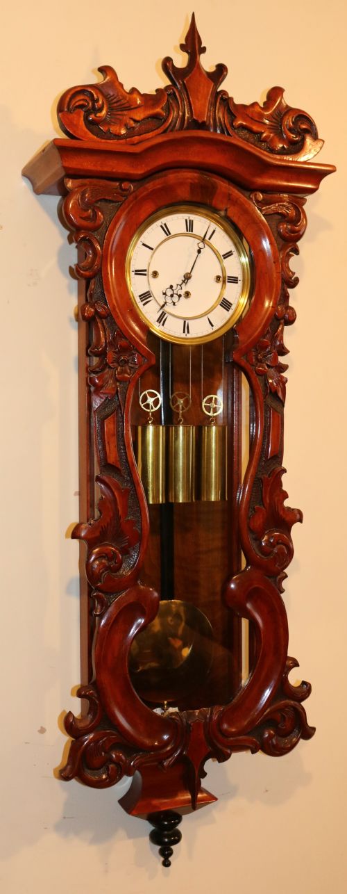 fig 1. An antique carved walnut Vienna wall clock featuring a glazed case, white enamel dial with Roman numerals, carved scrollwork pediment, and three brass weights.