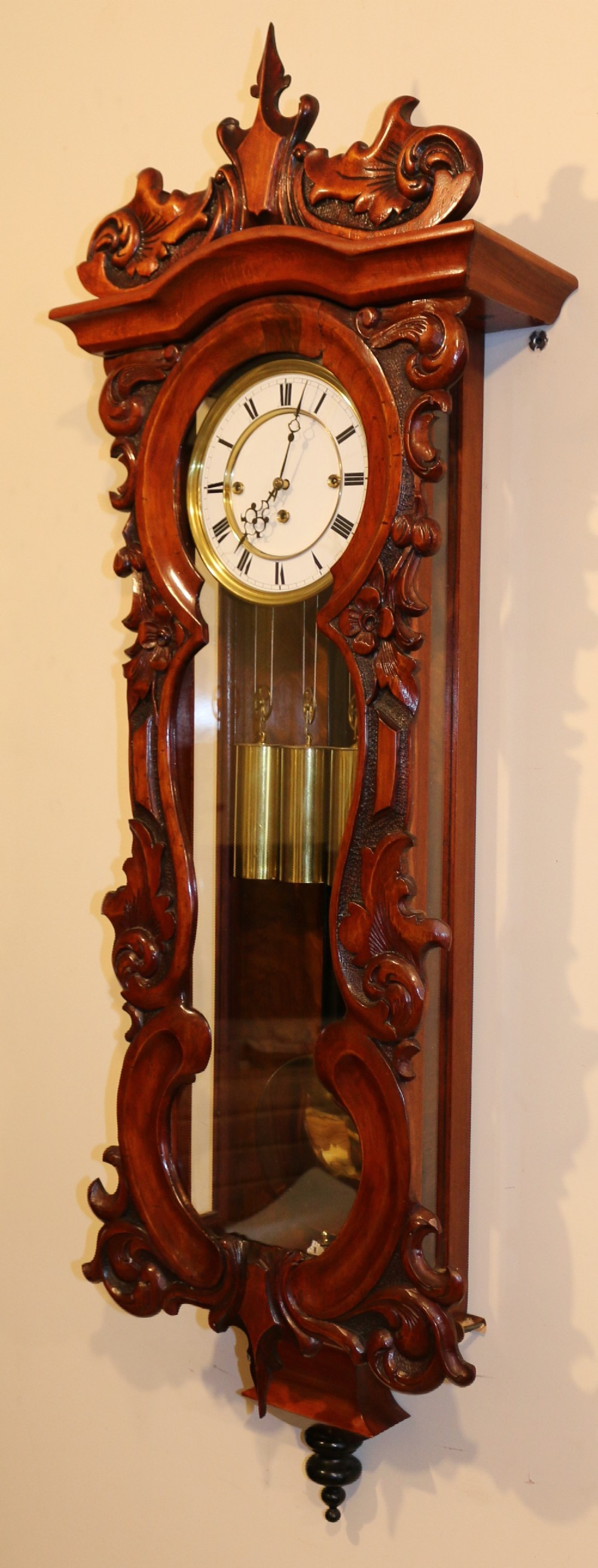 An antique carved walnut Vienna wall clock featuring a glazed case, white enamel dial with Roman numerals, carved scrollwork pediment, and three brass weights.