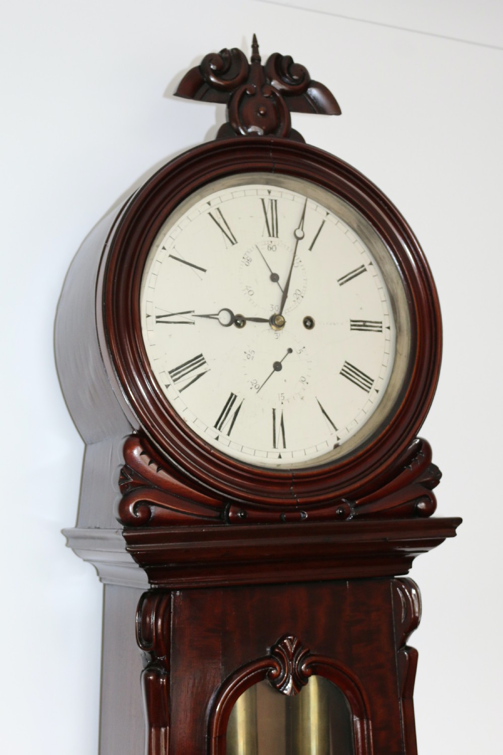 A tall flame mahogany Scottish regulator longcase clock featuring a circular white painted dial, carved top scrollwork, and a glazed door showing brass weights. View III