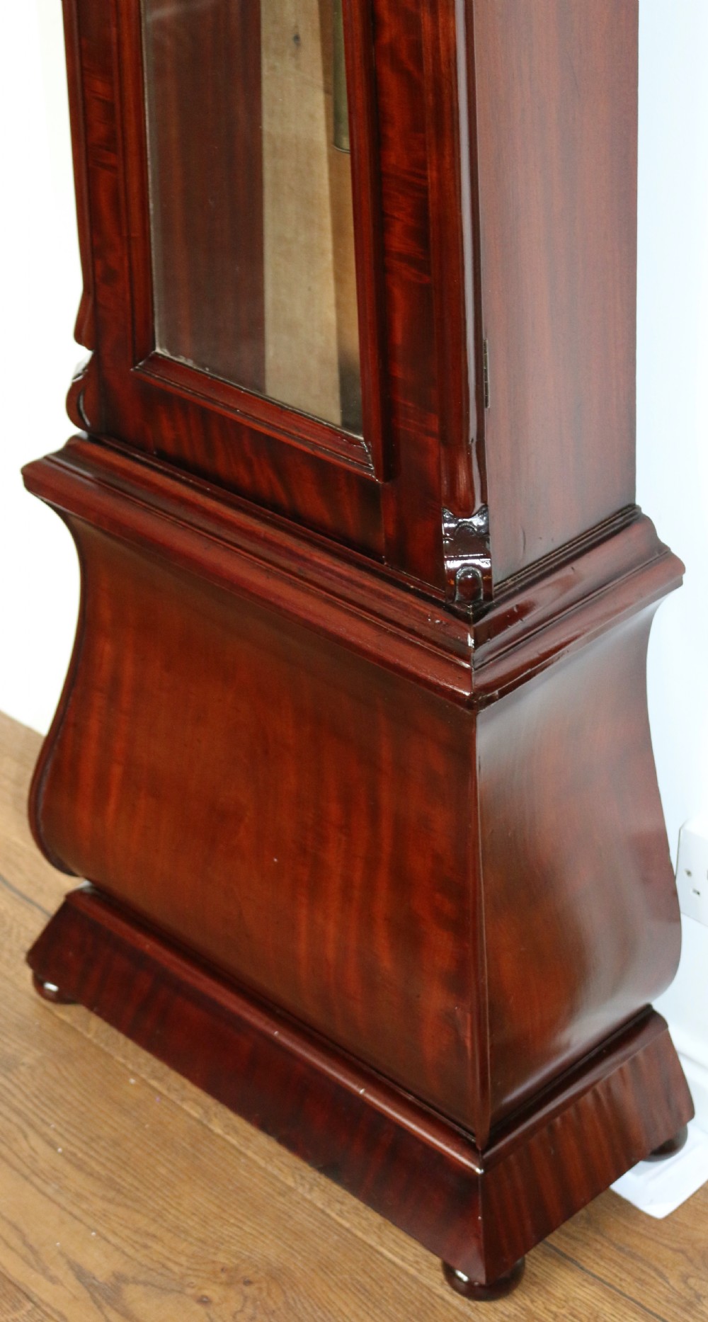 A tall flame mahogany Scottish regulator longcase clock featuring a circular white painted dial, carved top scrollwork, and a glazed door showing brass weights. View IV