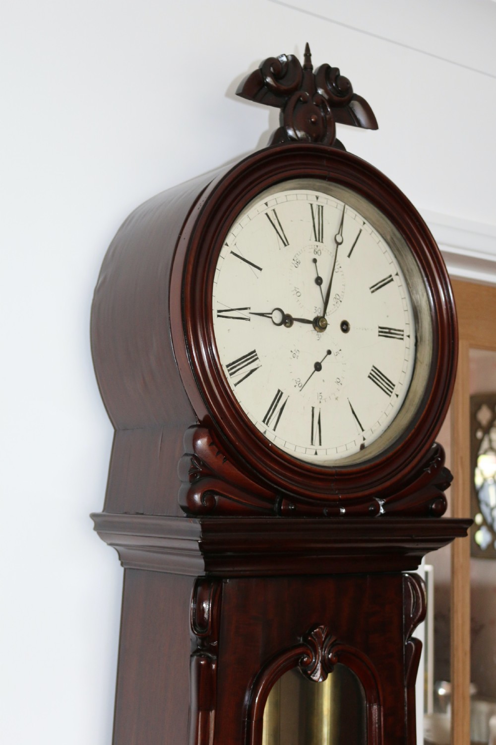 A tall flame mahogany Scottish regulator longcase clock featuring a circular white painted dial, carved top scrollwork, and a glazed door showing brass weights. View VI