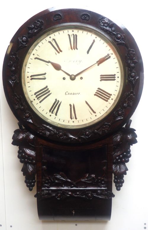 fig 1. An antique Victorian carved dark mahogany wall clock featuring an off-white dial with Roman numerals marked Berry Cardiff, framed by thistle and vine carvings.