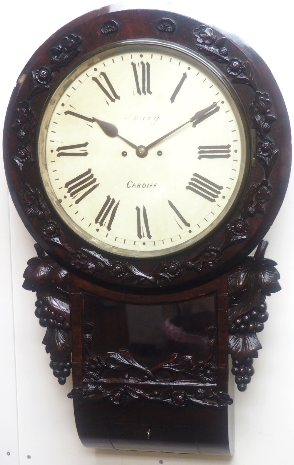 An antique Victorian carved dark mahogany wall clock featuring an off-white dial with Roman numerals marked Berry Cardiff, framed by thistle and vine carvings.