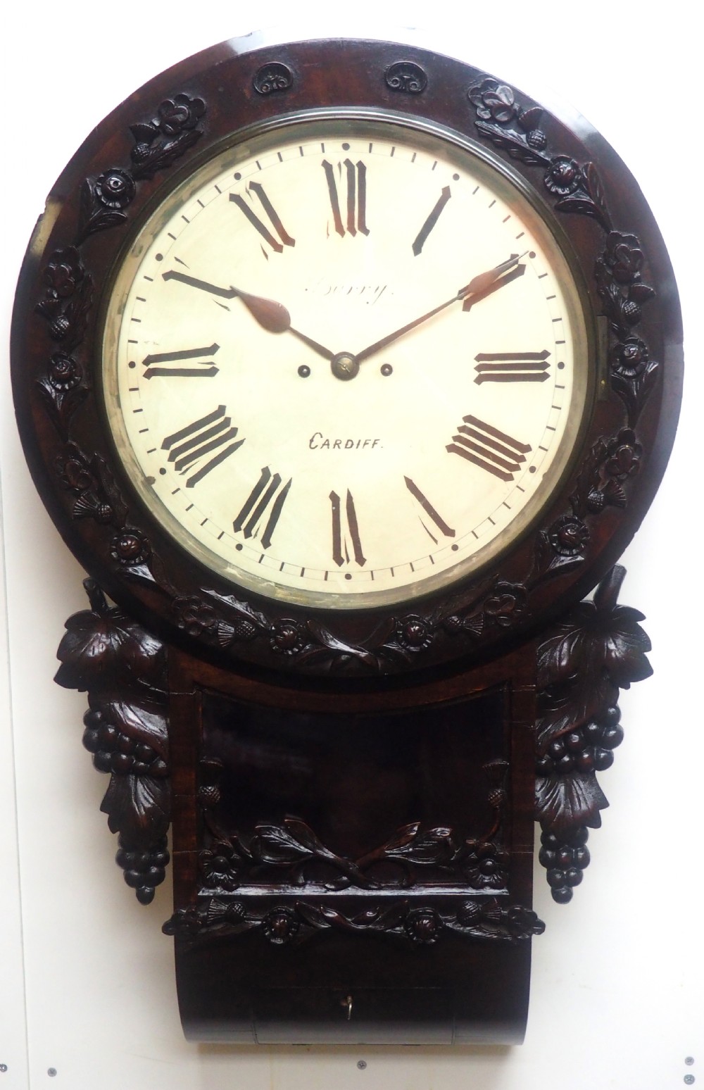An antique Victorian carved dark mahogany wall clock featuring an off-white dial with Roman numerals marked Berry Cardiff, framed by thistle and vine carvings. View II